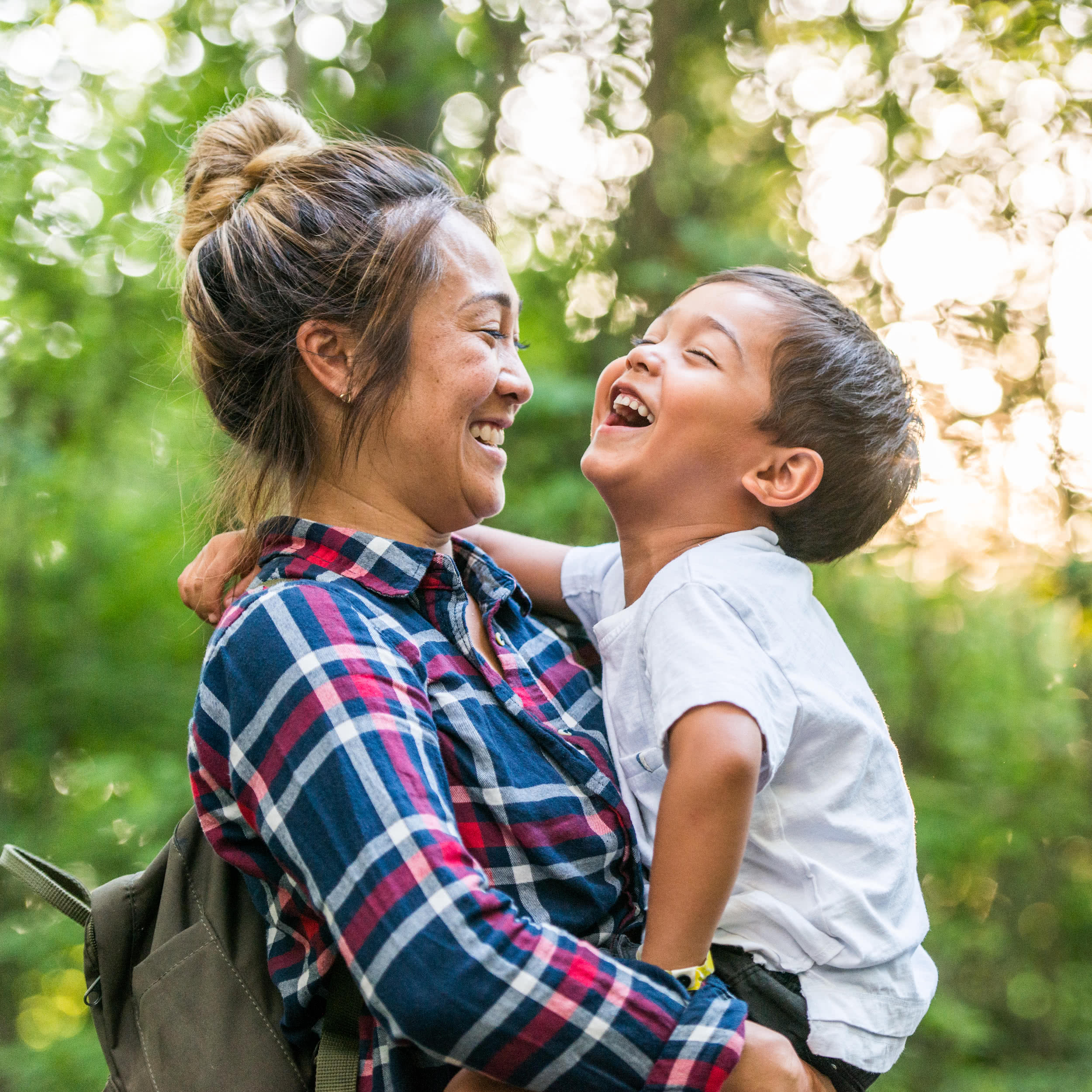 Woman with backpack laughing together with a child as she carries them on a walk in the forest