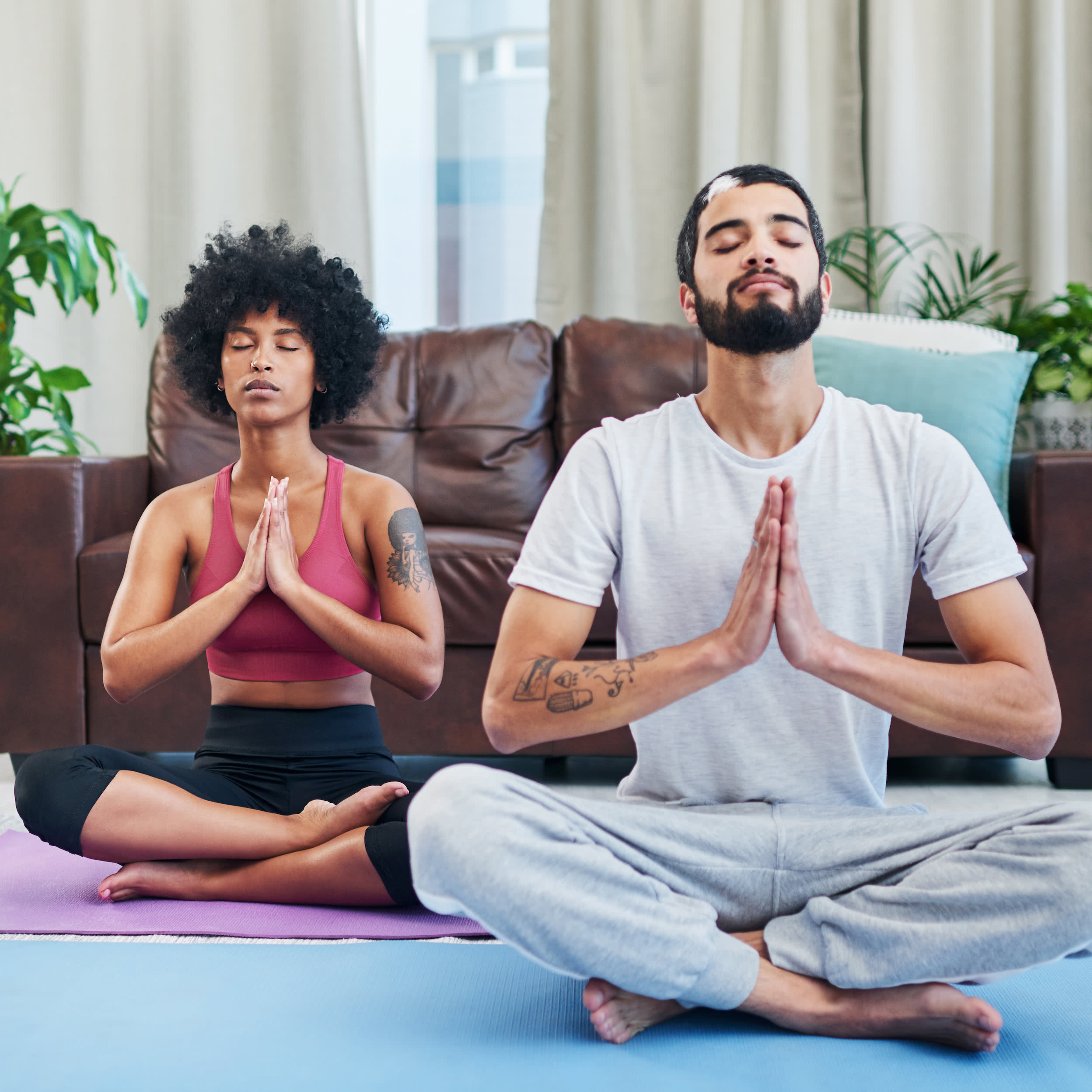 Two people sitting on yoga mats on the floor of a living room, each doing the same meditation pose
